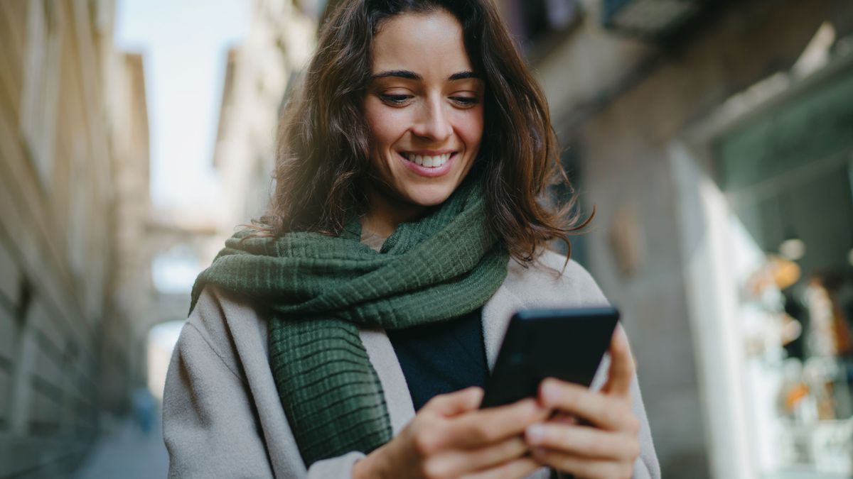 Portrait of young smiling dark-haired woman wearing light-colored coat and green scarf, walking city streets and using smartphone to interact with her friends. Good vibes. Modern technologies concept