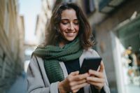 Portrait of young smiling dark-haired woman wearing light-colored coat and green scarf, walking city streets and using smartphone to interact with her friends. Good vibes. Modern technologies concept