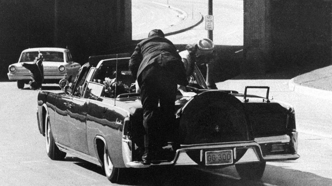 Präsident John F. Kennedy sackt auf den Rücksitz der Präsidentenlimousine zusammen, während diese die Elm Street in Richtung des Stemmons-Freeway-Überführungs in Dallas, Texas, entlangrast, nachdem er am 22. November 1963 tödlich angeschossen wurde. First Lady Jacqueline Kennedy beugt sich über den Präsidenten, während Secret Service Agent Clint Hill sie zurück auf ihren Sitz drängt.