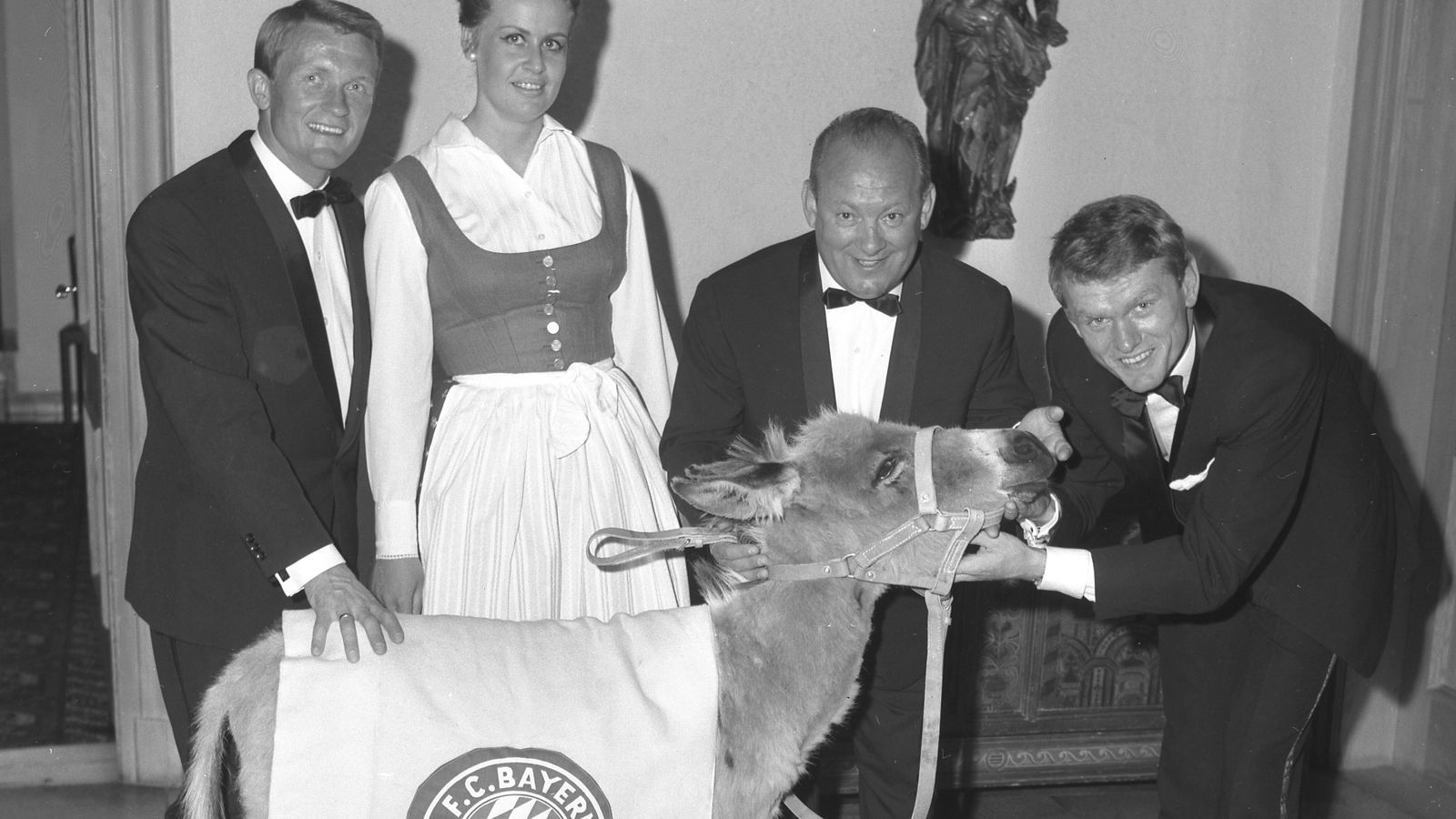 
                <strong>FC Bayern München: Die Maskottchen im Wandel der Zeit</strong><br>
                Dieses Foto ist das älteste überlieferte Dokument eines Bayern-Maskottchens. Am 18. Juni 1967 übergibt Hotelchefin Karin Rauh (geb. Bachmaier) im Hotel Bachmaier am Tegernsee den Esel Sepp an Torwart Sepp Maier (r.), nach dem der Esel auch benannt ist. Kapitän Werner Olk (l.) und Trainer Tschik Cajkovski schauen begeistert zu. Im Hotel Bachmaier begossen die Bayern ihre ersten großen Erfolge, die Hotelchefin revanchierte sich mit dem lebenden Maskottchen.
              