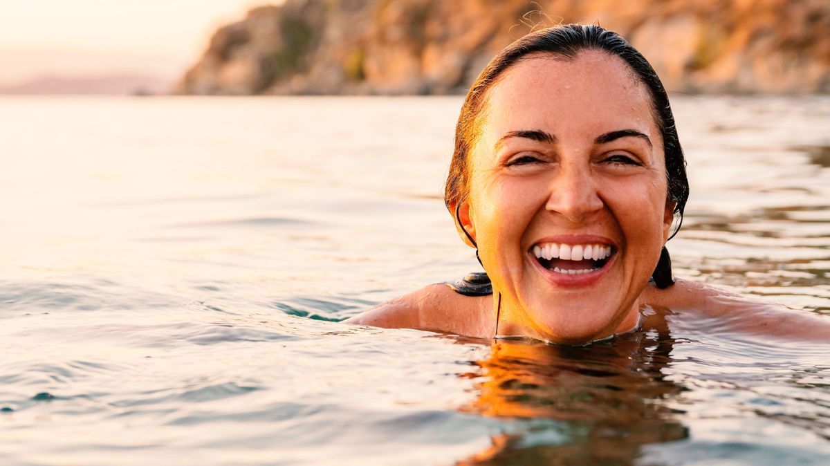 Caucasian woman laughing and showing her white teeth while swimming in sea at sunset