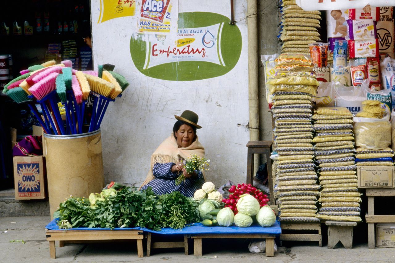 Diese Frau in traditioneller Kleidung mit Schultertuch und Hut verkauft frisches Obst und Gemüse an einem Straßenstand - ein typisches Bild.