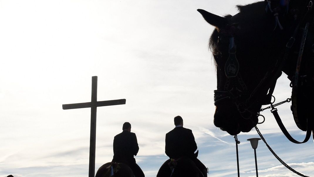 Männer reiten bei der traditionellen Leonhardifahrt an einem Kreuz vorbei. Bei einer traditionellen Prozession in Schwaben bricht ein Teil einer Kutsche. 