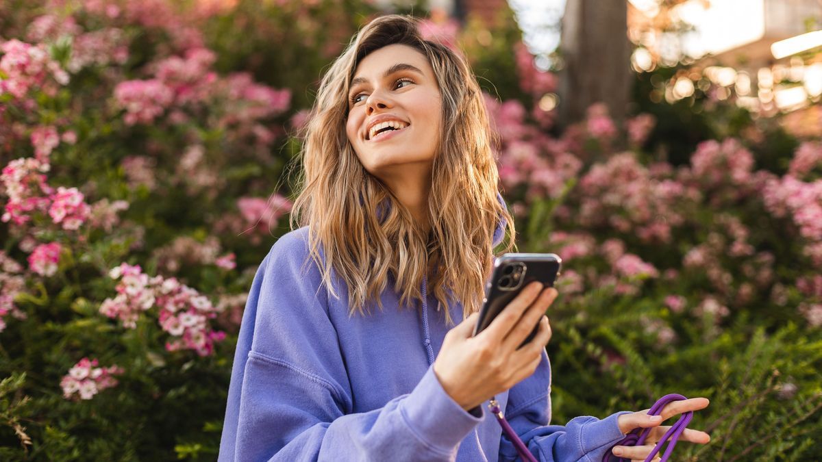 Attractive blonde curly woman wear purple hoody hold mobile phone and look at side, look happy and smiling. Amazing enjoy woman walk near blossom flowers.