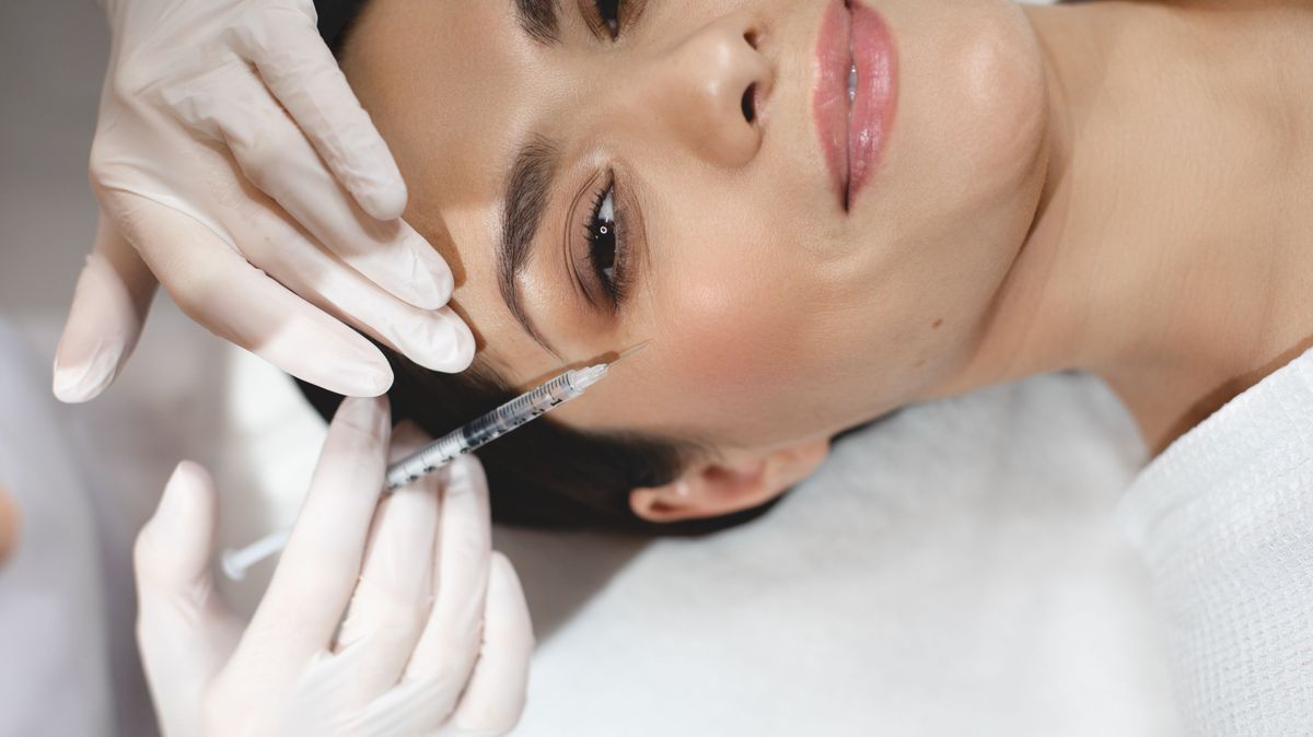 Cut view of happy positive young woman during botox injection process. Hands in white gloves holding syringe close to eye area and input some collagen under facial skin.