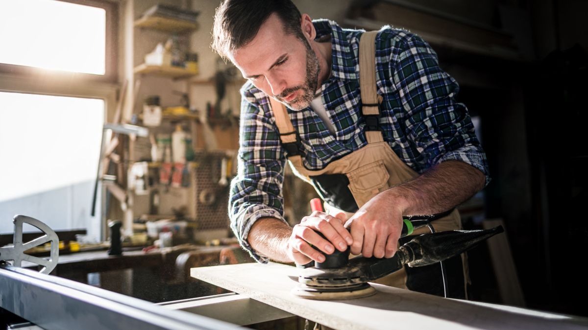 Ein Mann/Handwerker schleift ein Holzbrett mit einem Exzenterschleifer in einer Werkstatt ab
