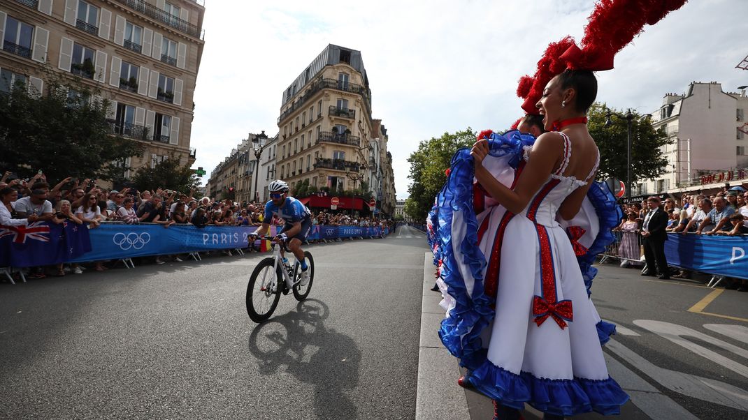 Olympia 2024: Männer, Tänzerinnen vor dem Moulin Rouge feuern die Radsportler an. 