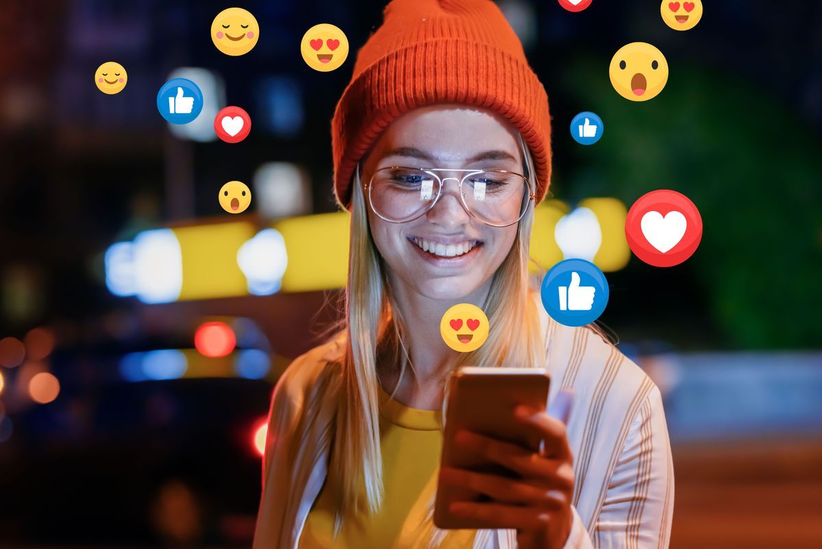 A beautiful young woman blogger, vlogger or influencer is receiving emoji and emoticon reactions in her mobile smart phone device while making a post, sharing or video logging on social media