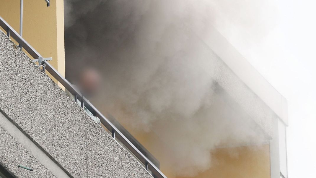Eine Person steht auf dem Balkon von einem Hochhaus, aus dem Rauch quillt.