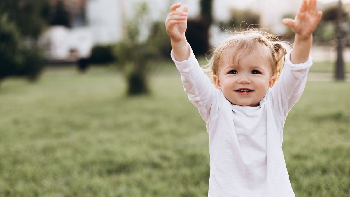 little baby girl with blond hair and two tails, wearing a white T-shirt standing on green grass in nature, smiling