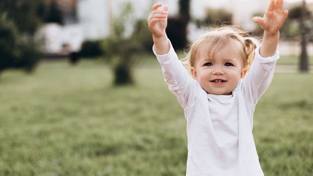 little baby girl with blond hair and two tails, wearing a white T-shirt standing on green grass in nature, smiling