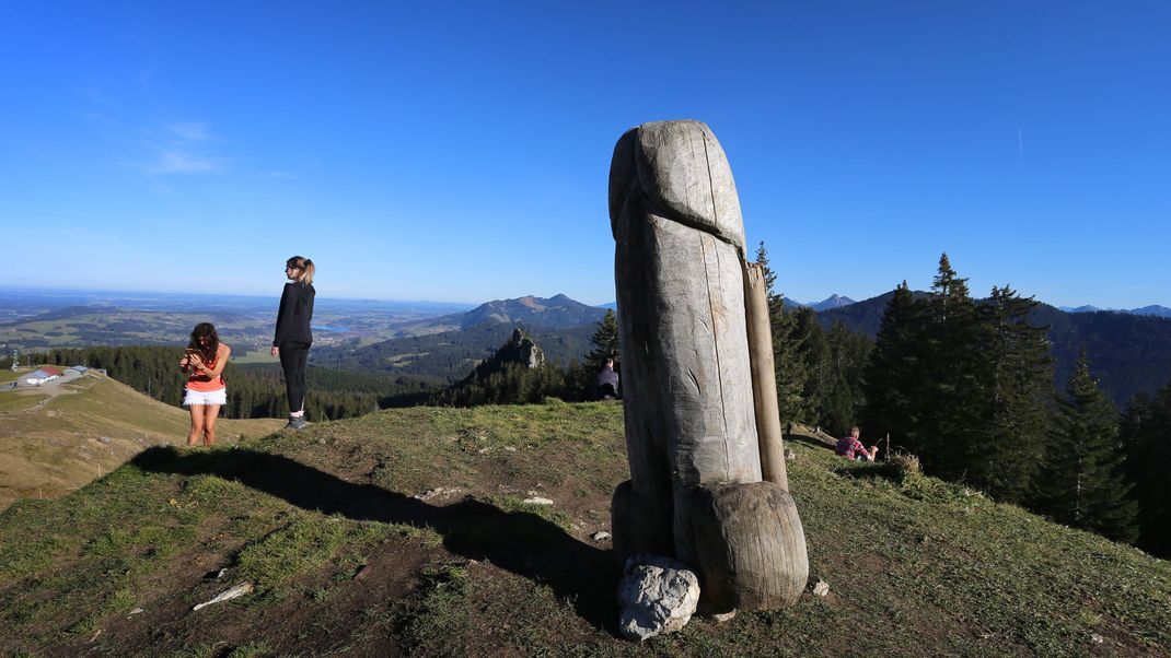 Wanderer genießen neben der zwei Meter hohen Penis-Skulptur aus Holz auf dem Grat des Grünten den Ausblick. 