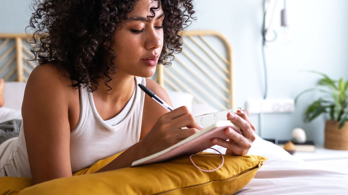Pensive young multiracial latina woman lying down on bed with serious expression writing on journal in cozy bedroom
