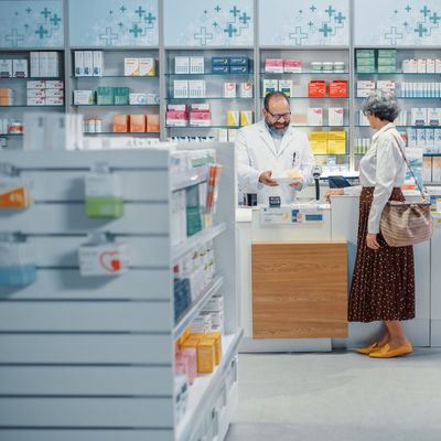 Pharmacy Drugstore Checkout Cashier Counter: Senior Woman Buying Prescription Medicine, Drugs, Vitamins and Talks to Beautiful Pharmacist Cashier, Asking Recomendations. Store with Health Care Goods