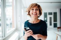 Cheerful businesswoman holding mobile phone while standing in office