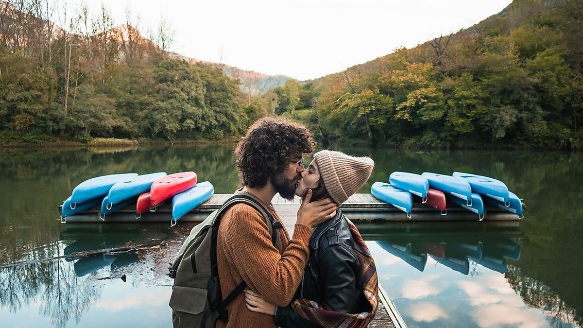 Side view young loving couple in warm wear kissing tenderly while standing on lake wooden quay against lush abundant forested hills in reservoir Valdemurio