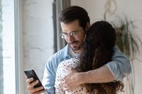 Dishonest young man using phone while cuddling wife.