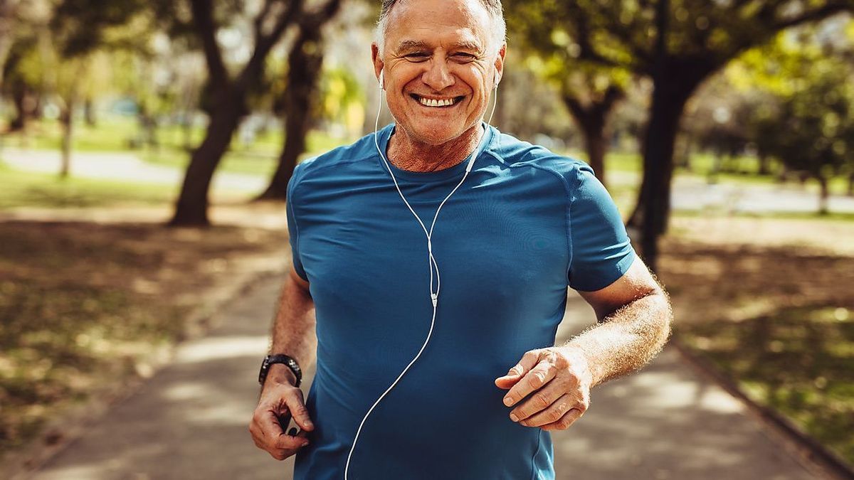 Porträt eines älteren Mannes in Sportbekleidung, der in einem Park läuft. Nahaufnahme eines lächelnden Mannes, der beim Laufen Musik hört und Kopfhörer verwendet.