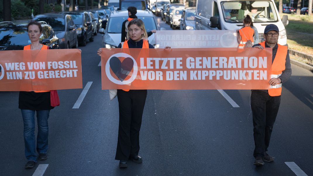 Laufen statt kleben: Aktivist:innen der "Letzten Generation" gehen mit Bannern vor dem Verkehr in Berlin her und zwingen diesen nur noch in Schrittgeschwindigkeit zu fahren.