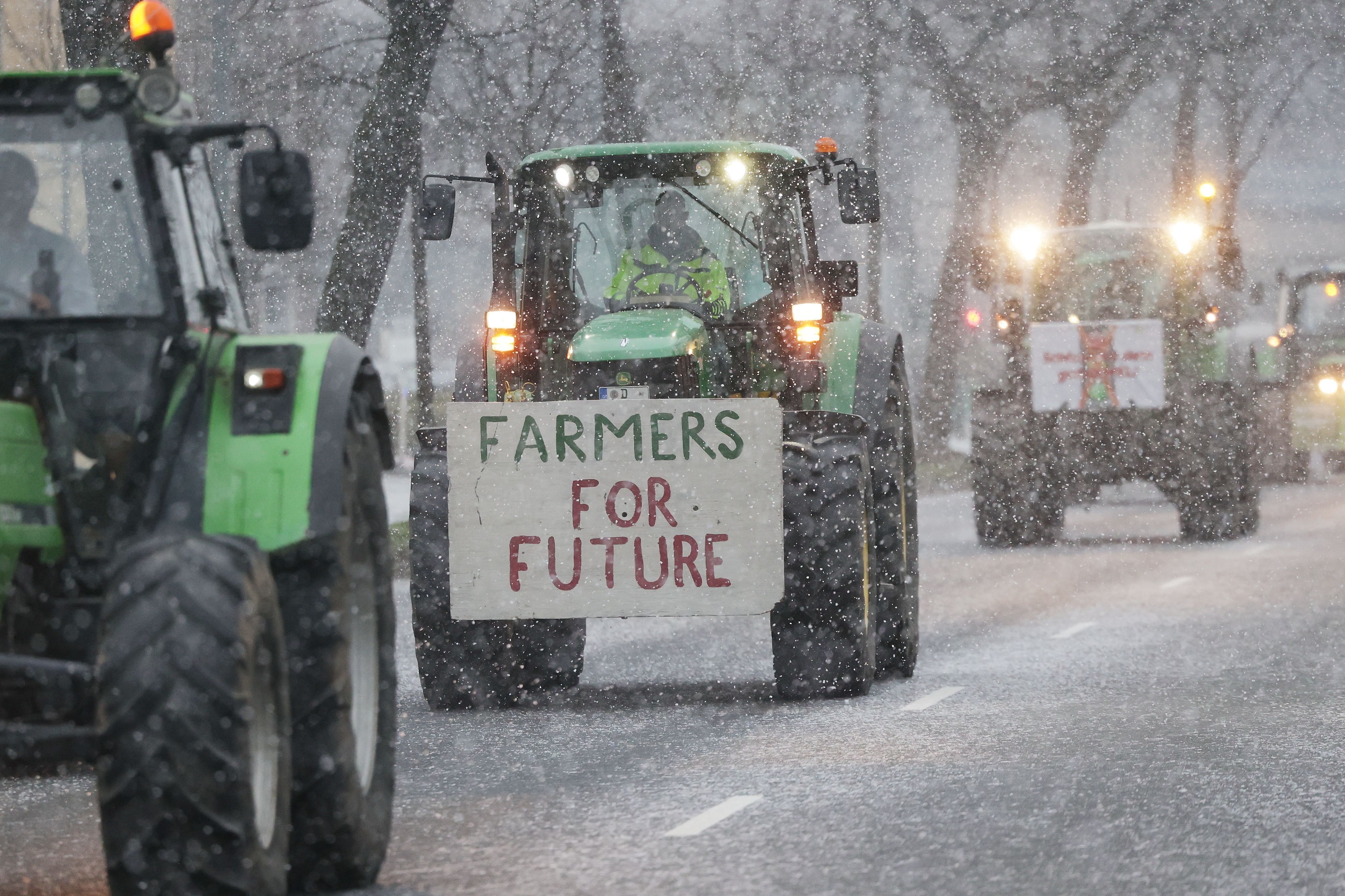 Im Rahmen der Protest-Aktionswoche der Landwirt:innen fahren die Traktoren auch über eine schneebedeckte Straße in Düsseldorf. Auf einem Fahrzeug haben Landwirte dabei ein Schild mit der Aufschrift "Farmers For Future" befestigt.