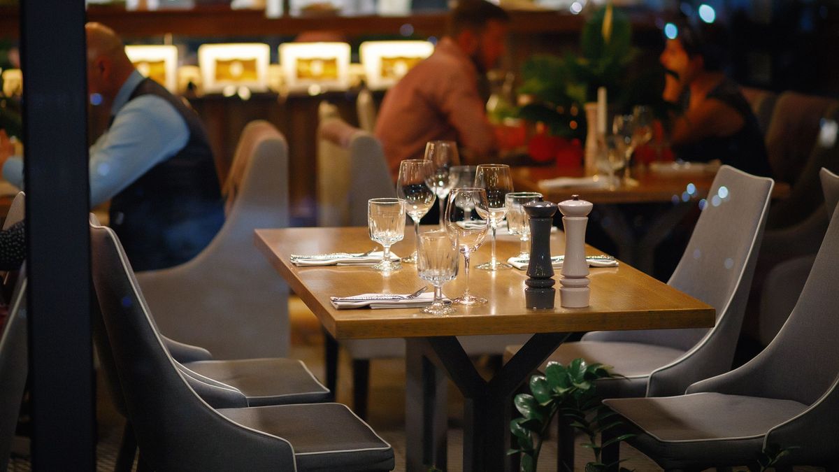 empty served table in an expensive restaurant for four people with glasses, plates and appliances in the background visitors and 3 cooks in white. evening interior of a popular cafe