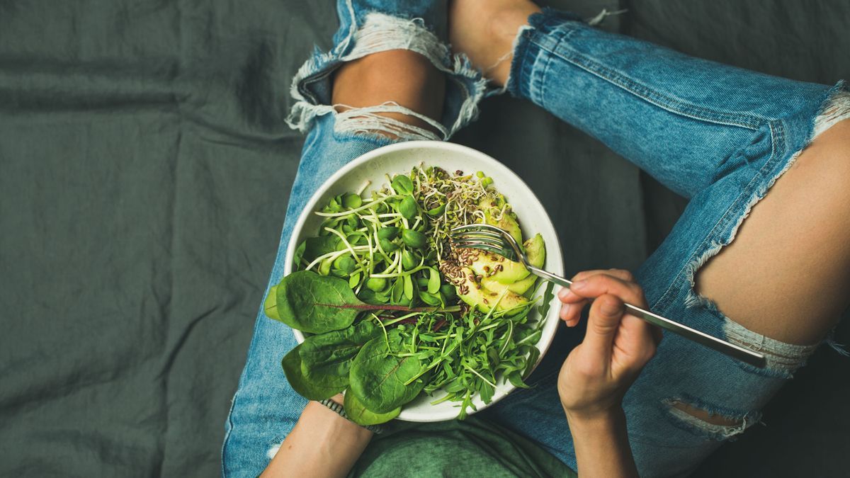 Green vegan breakfast meal in bowl with spinach, arugula, avocado, seeds and sprouts. Girl in jeans holding fork with knees and hands visible, top view, copy space. Clean eating, vegan food concept