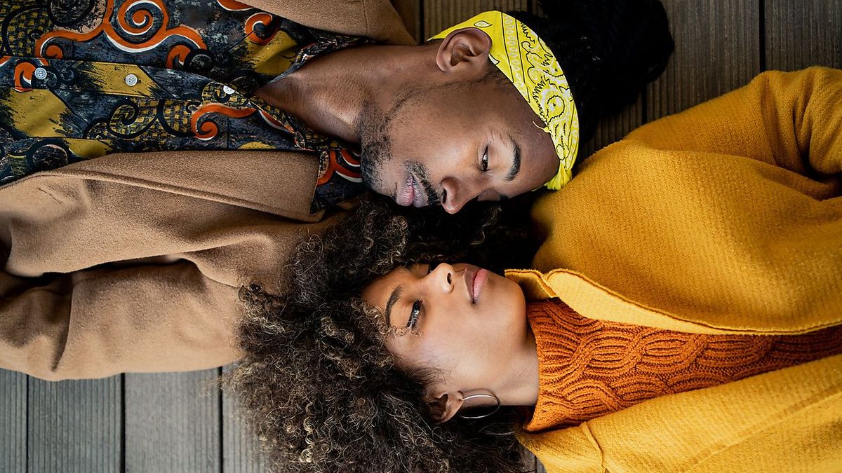 Top view of trendy African American man and woman in bright coat lying opposite with face to face looking at each other