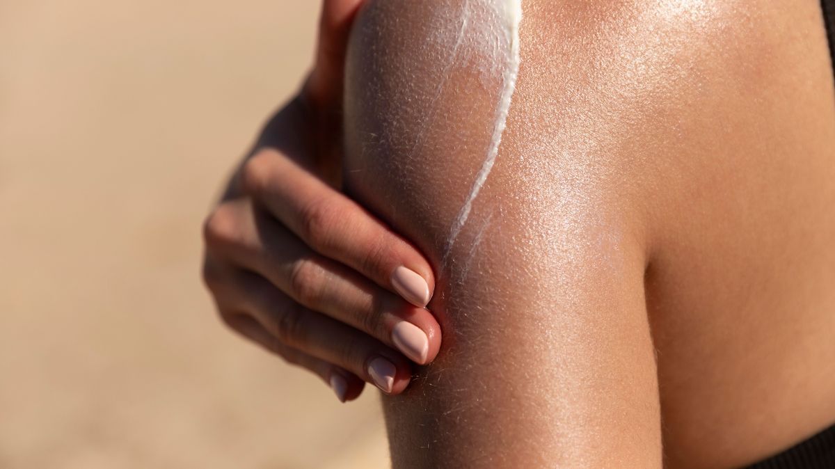 Young woman applying sun cream or sunscreen on her tanned shoulder to protect her skin from the sun. Shot on a sunny day with blurry sand in the background
