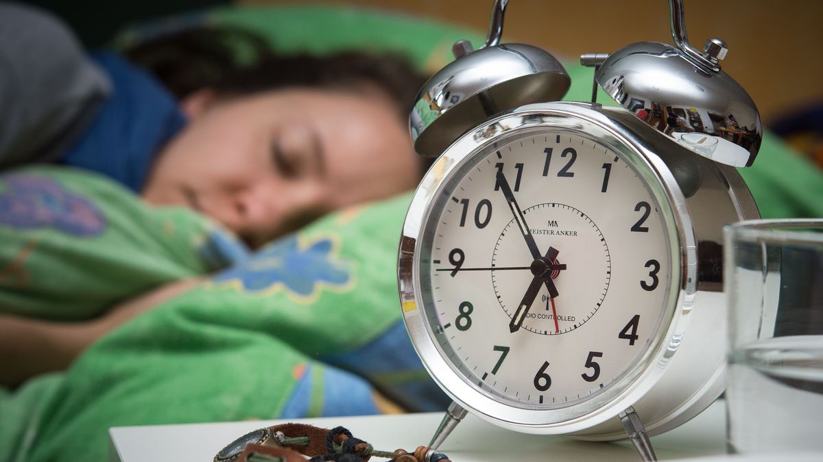 Eine Frau schläft im Bett, während ein Wecker, Wasserglas, Schmuck und ein Buch vor ihr auf dem Nachttischränkchen zu sehen sind. 