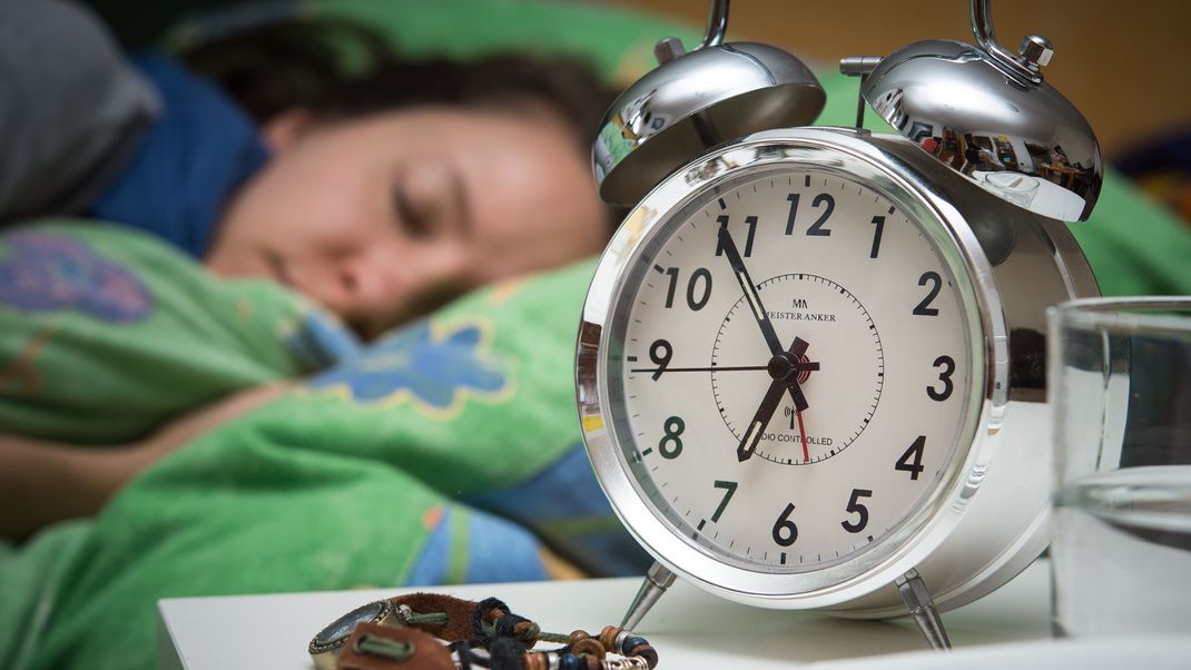 Eine Frau schläft im Bett, während ein Wecker, Wasserglas, Schmuck und ein Buch vor ihr auf dem Nachttischränkchen zu sehen sind. 