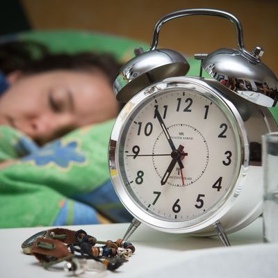 Eine Frau schläft im Bett, während ein Wecker, Wasserglas, Schmuck und ein Buch vor ihr auf dem Nachttischränkchen zu sehen sind. 