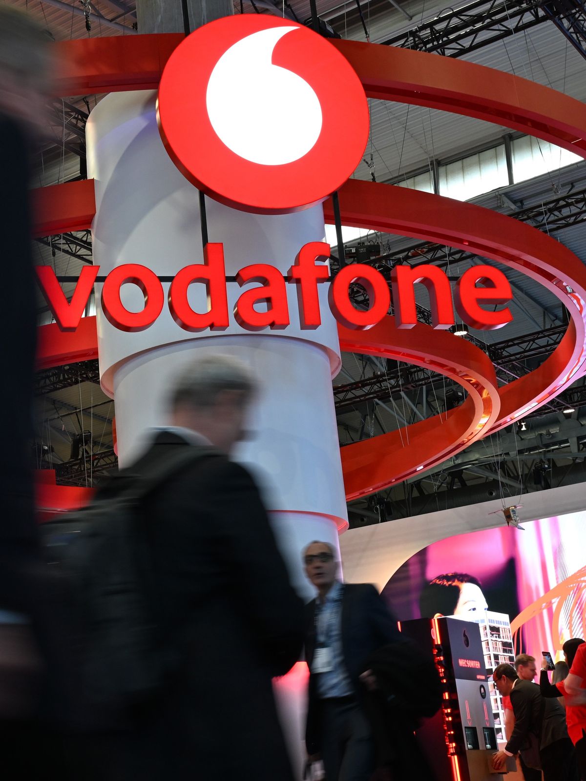 3. März 2025, Spanien, Barcelona: Menschen gehen auf der Mobilfunk-Messe Mobile World Congress (MWC) am Stand des Telekommunikationskonzerns Vodafone vorbe