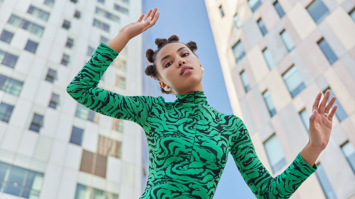 Below view of serious stylish teenage girl with makeup keeps arms raised dressed in comfortable clothes poses against modern skyscrapers against blue sky stands at downtown. Urban lifestyle concept