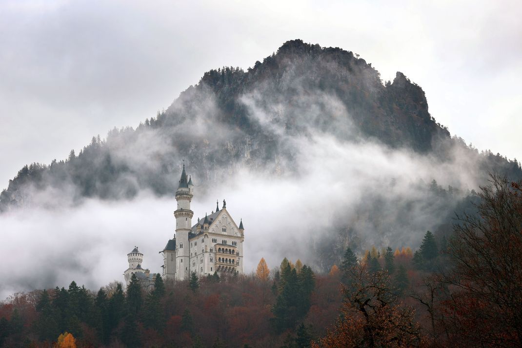 Wolkenverhangen steht das Schloss Neuschwanstein in der herbstlichen Landschaft des Königswinkels.