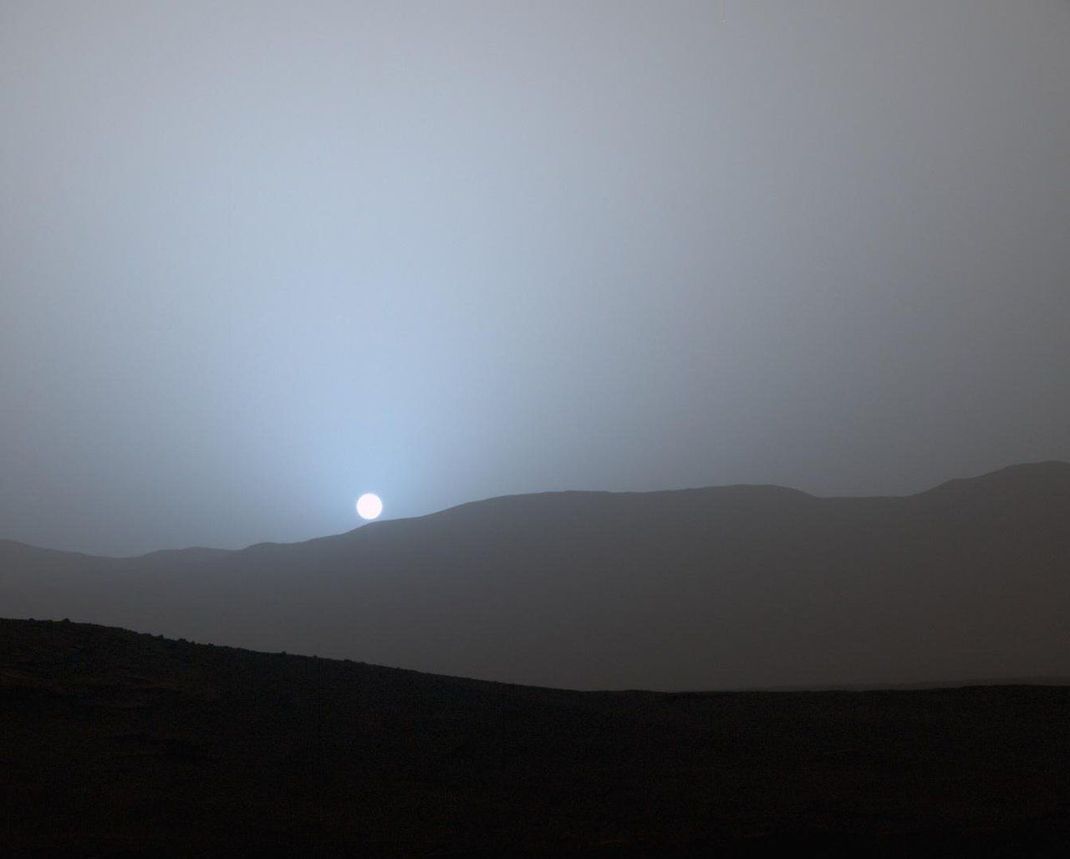 Sonnenuntergang auf dem Mars, aus der Sicht des "Curiosity"-Rovers. Da der Planet im Schnitt etwa 1,5 Mal weiter von der Sonne entfernt ist als die Erde, fällt er deutlich weniger romantisch aus als zum Beispiel auf Sylt. Der Staub in der Marsluft filtert den roten Lichtanteil weitgehend raus, daher wird der Marshimmel abends blaugrau statt rot.