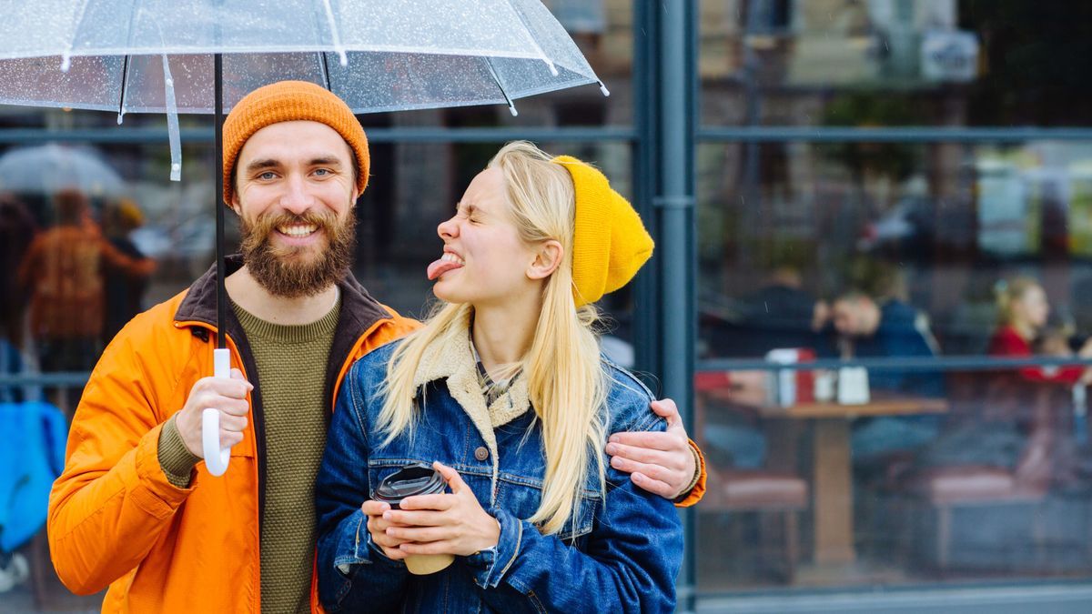 girl showing tongue making boyfriend laugh, joking, grimacing teasing while hugging under an transparent umbrella in rainy weather