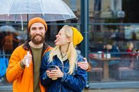 girl showing tongue making boyfriend laugh, joking, grimacing teasing while hugging under an transparent umbrella in rainy weather