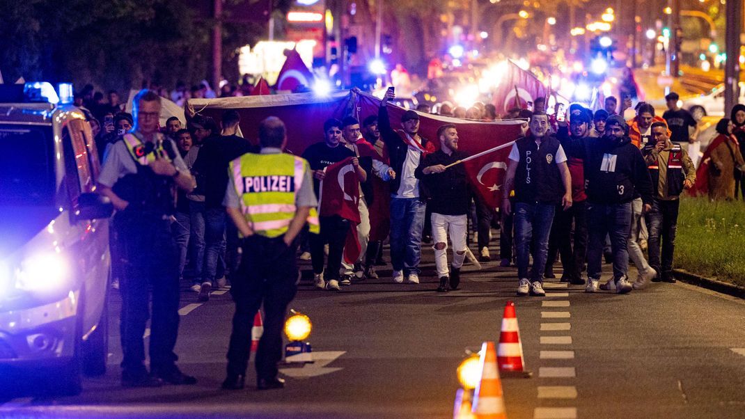 Hunderte Anhänger des amtierenden türkischen Präsidenten Erdogan laufen in Duisburg mit Flaggen über eine Straße. Die Polizei ist am späten Abend mit starken Kräften vor Ort.