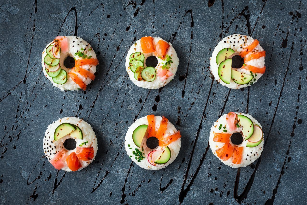 Jetzt geht‘s rund! Es gibt nicht nur süße Kringel, sondern mittlerweile auch Sushi-Donuts. Wie man die herstellt? Der Reis wird zunächst in eine Ringform gepresst und anschließend beispielsweise mit Lachs, Avocado, Gurke, Wasabi und Sesam dekoriert. 