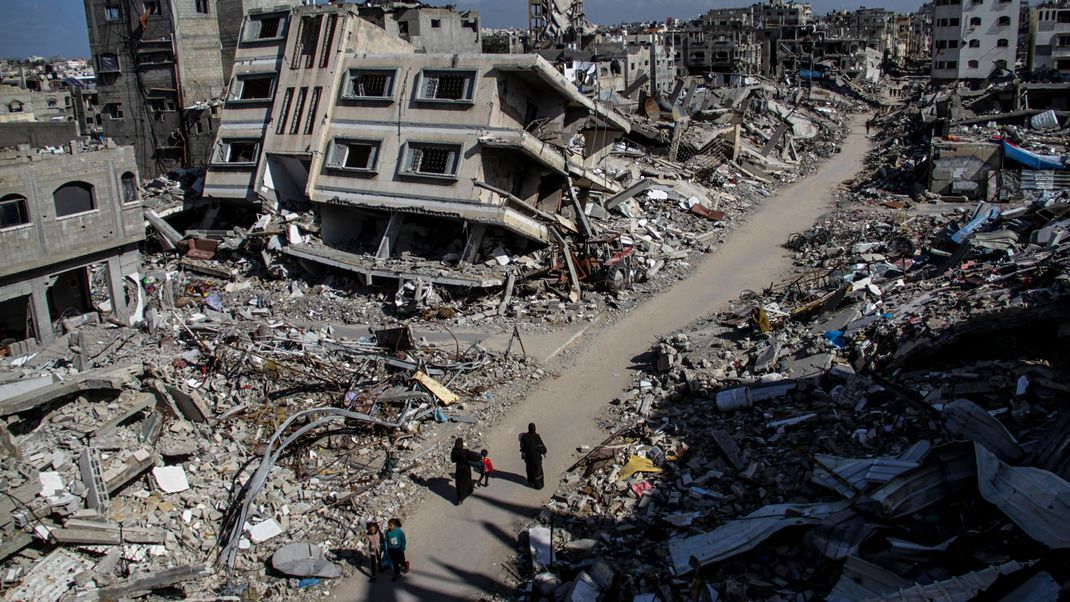 22.03.2024, Palästinensische Gebiete, Dschabalia: Menschen gehen an den Ruinen von Gebäuden vorbei, die durch israelischen Luftangriff im Flüchtlingslager Dschabalia zerstört wurden.
