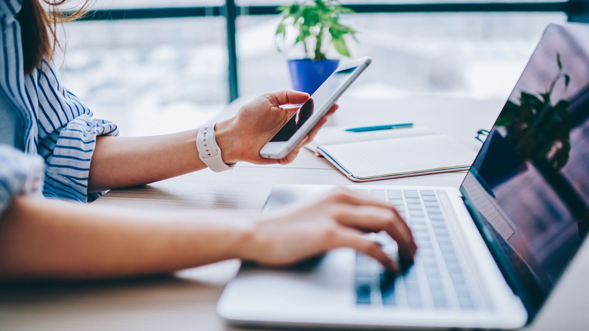 Eine Frau sitzt am Laptop und hält ihr Handy in der Hand.