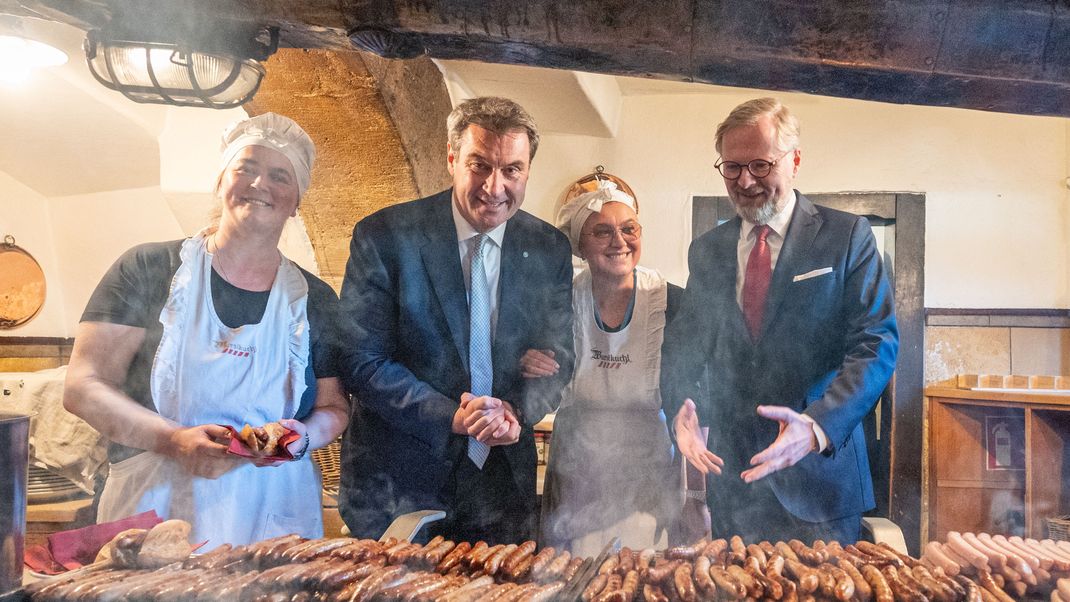 Petr Fiala, (r) Ministerpräsident der Tschechischen Republik und Markus Söder (CSU) stehen in der historischen Wurstkuchl, eine Bratwurststube, hinter einem Grill mit Bratwürsten.