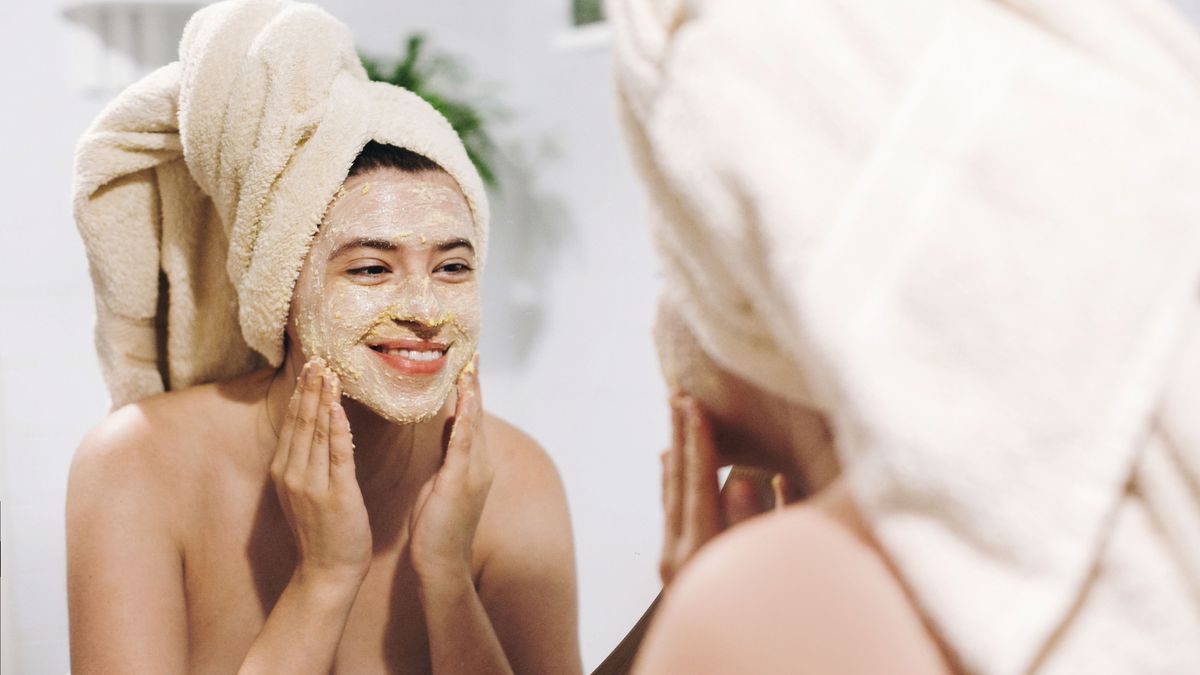 Skin Care concept. Young happy woman in towel making facial massage with organic face scrub and looking at mirror in stylish bathroom. Girl applying scrub cream, peeling and cleaning skin