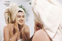 Skin Care concept. Young happy woman in towel making facial massage with organic face scrub and looking at mirror in stylish bathroom. Girl applying scrub cream, peeling and cleaning skin