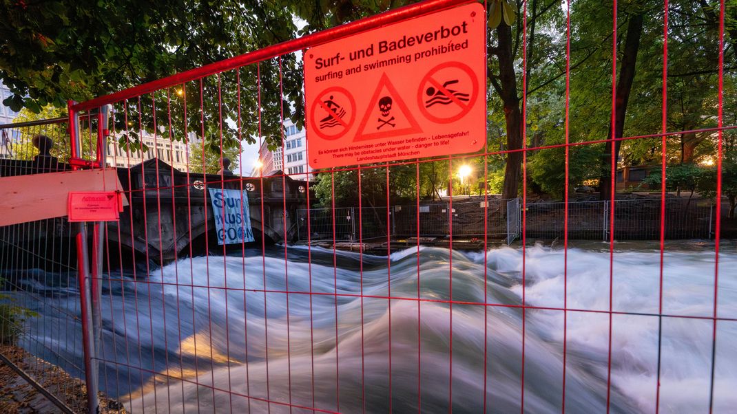 Ein Schild mit der Aufschrift „Surf- und Badeverbot“ hängt in den frühen Morgenstunden an einem Absperrzaun am Ufer der künstliche Eisbachwelle im Englischen Garten. 