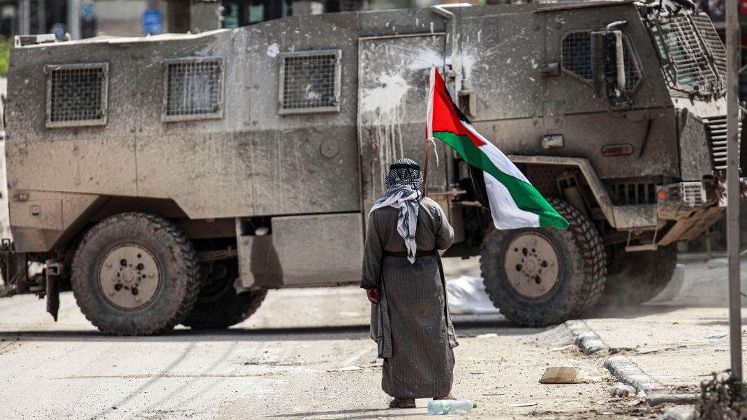 Ein älterer palästinensischer Mann steht mit einer palästinensischen Flagge in der Hand vor einem israelischen Militärfahrzeug.