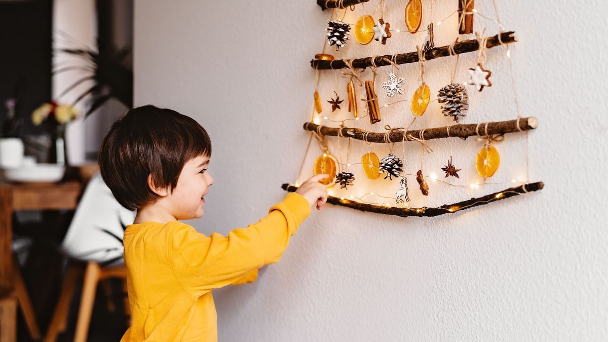 "Kleines Kind liegt neben einem selbstgemachten Weihnachtsbaum aus Zweigen und natürlichen Materialien, der an der Wand hängt. Nachhaltiges Weihnachten, kein Abfall, plastikfrei, umweltfreundlich."
