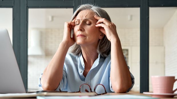 Ältere Frau sitzt mit Kopfschmerzen am Schreibtisch. 