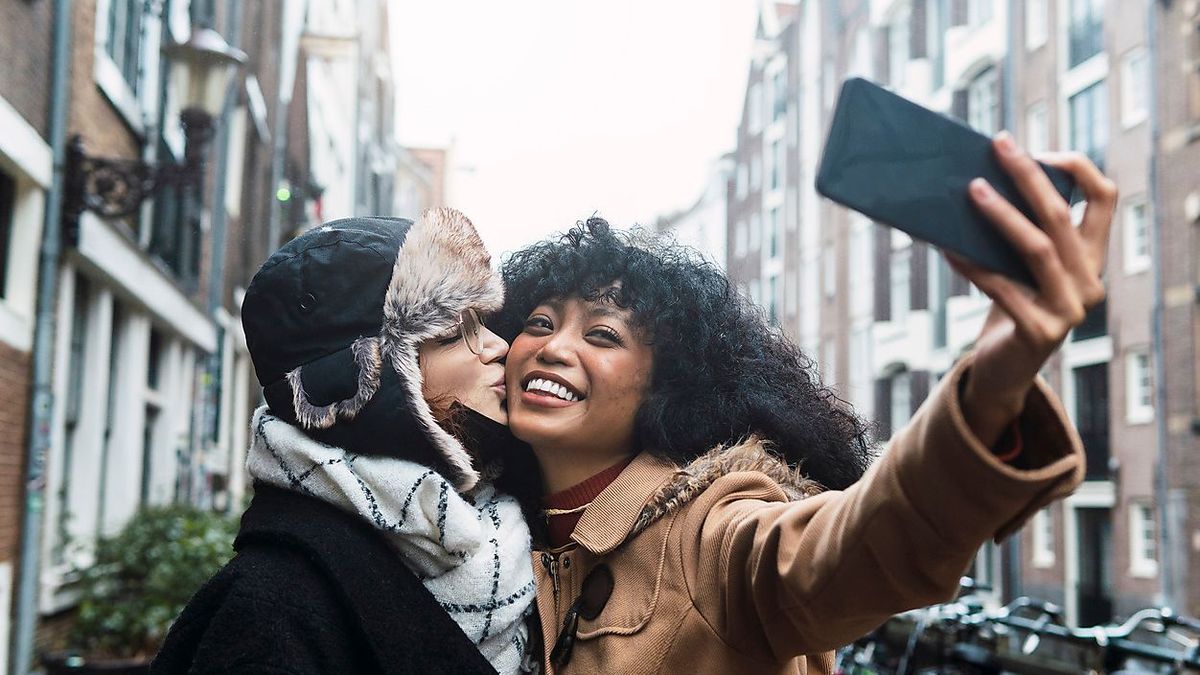 Happy young woman taking selfie with friend kissing on cheek in city