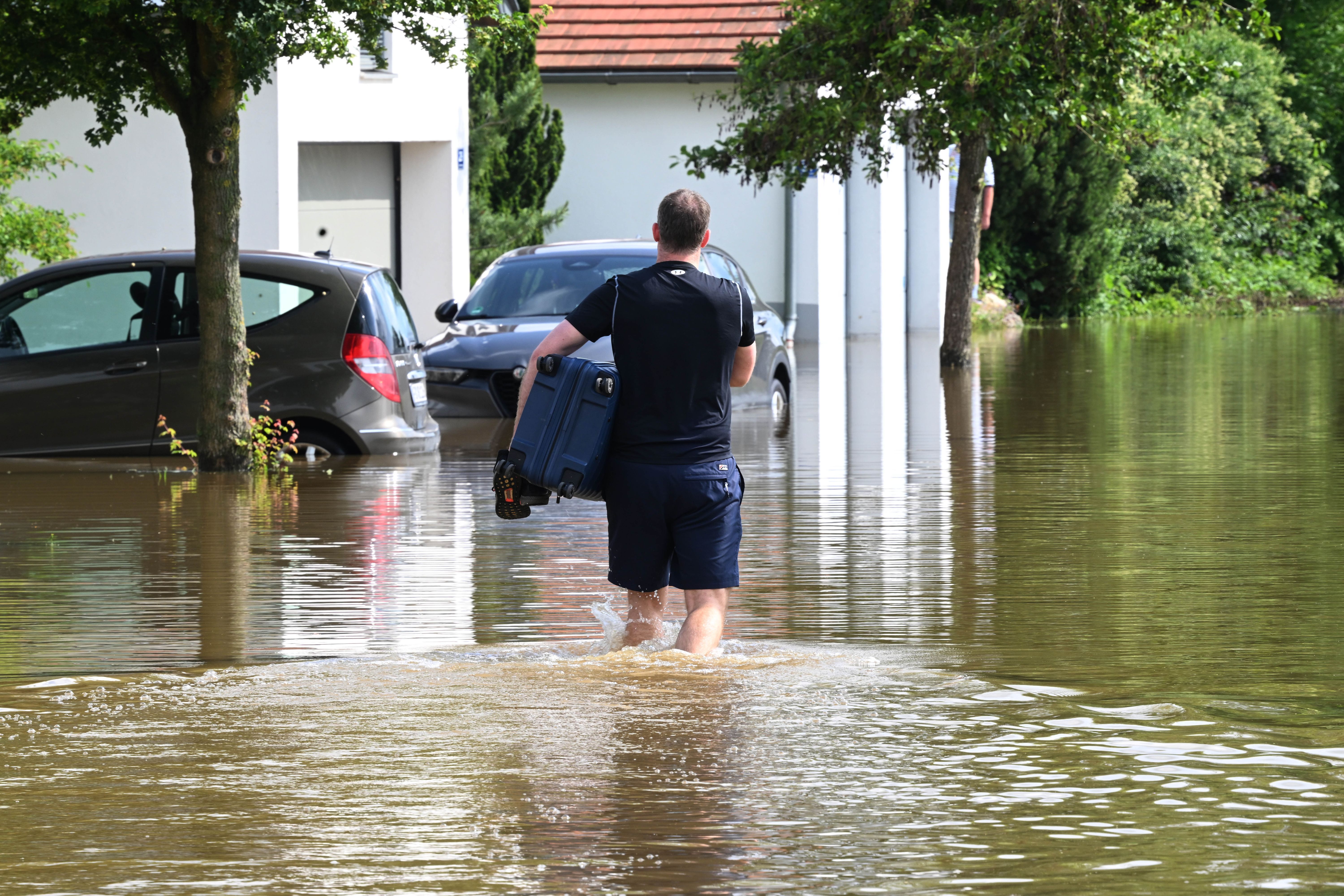 Ein Mann watet mit einem Koffer unter dem Arm durch eine überschwemmte Straße in einem Wohngebiet im bayerischen Farenzhausen.